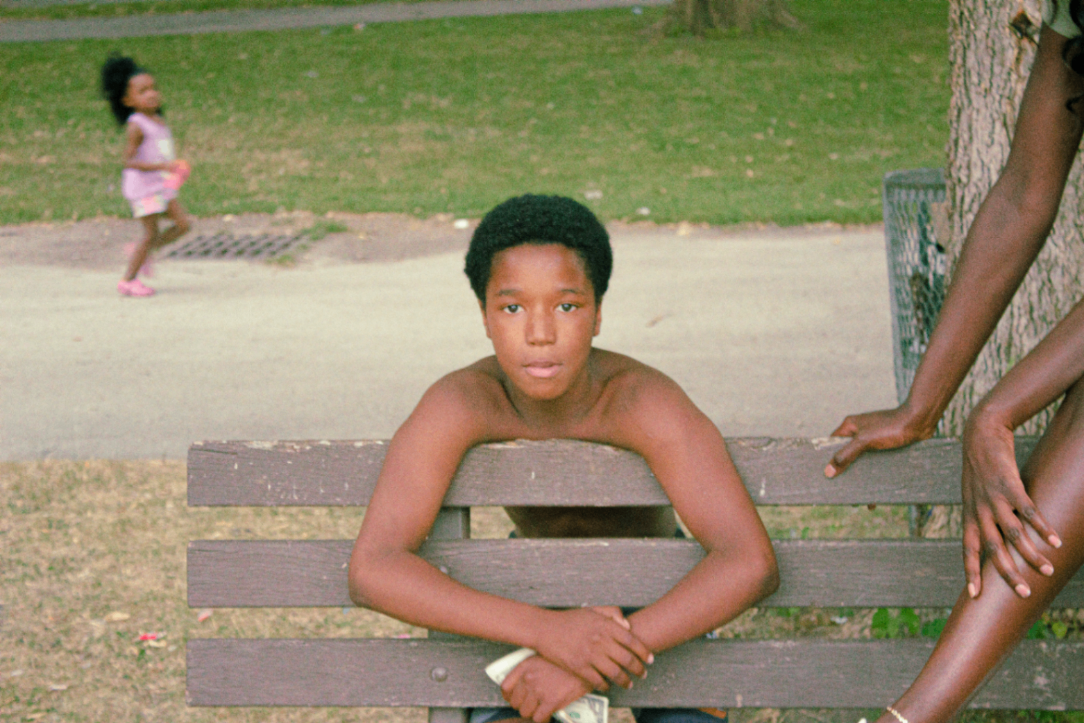 Young boy resting on a park bench with a child running past
