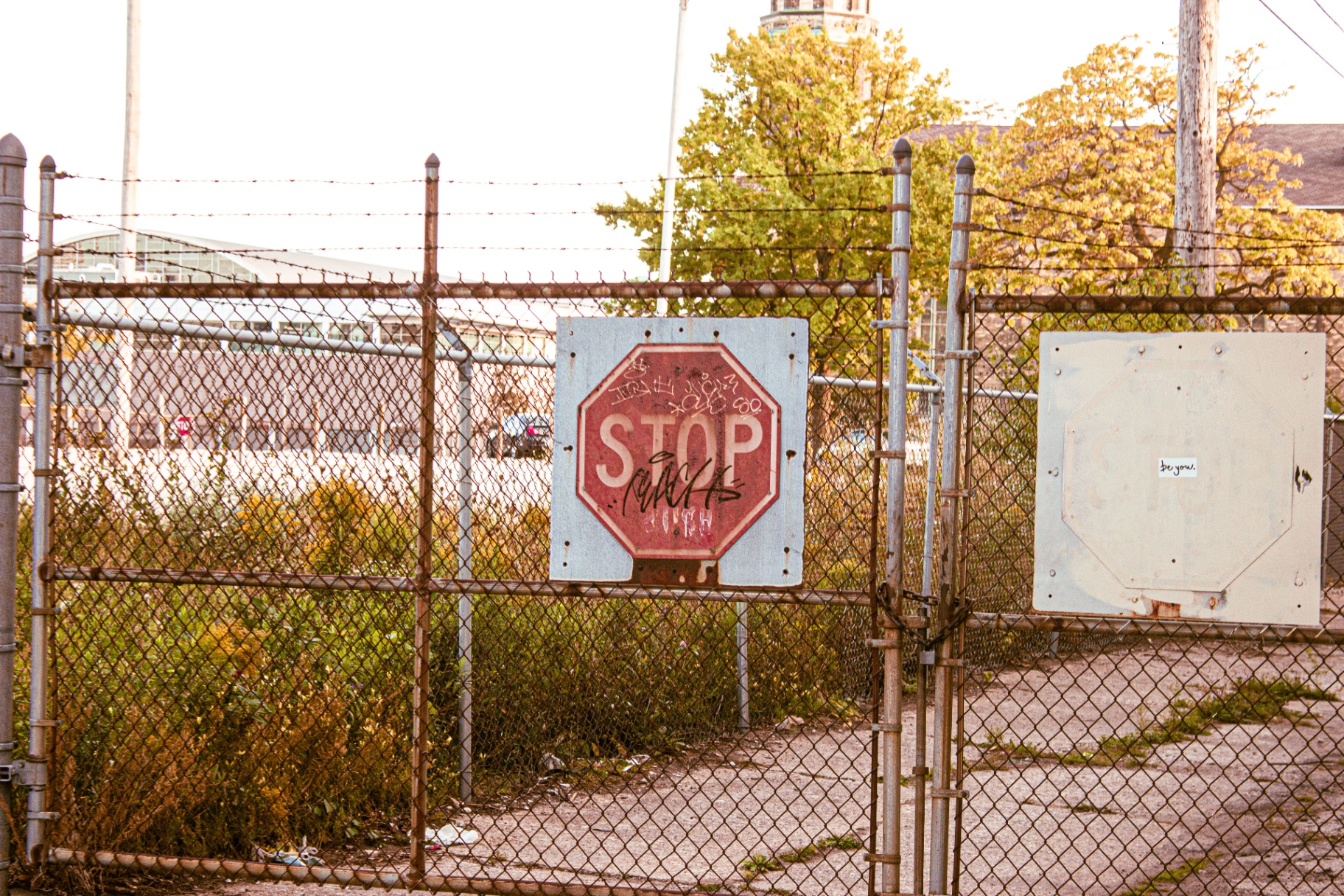 Graffitied stop sign on a chainlink gate with be you sticker beside it